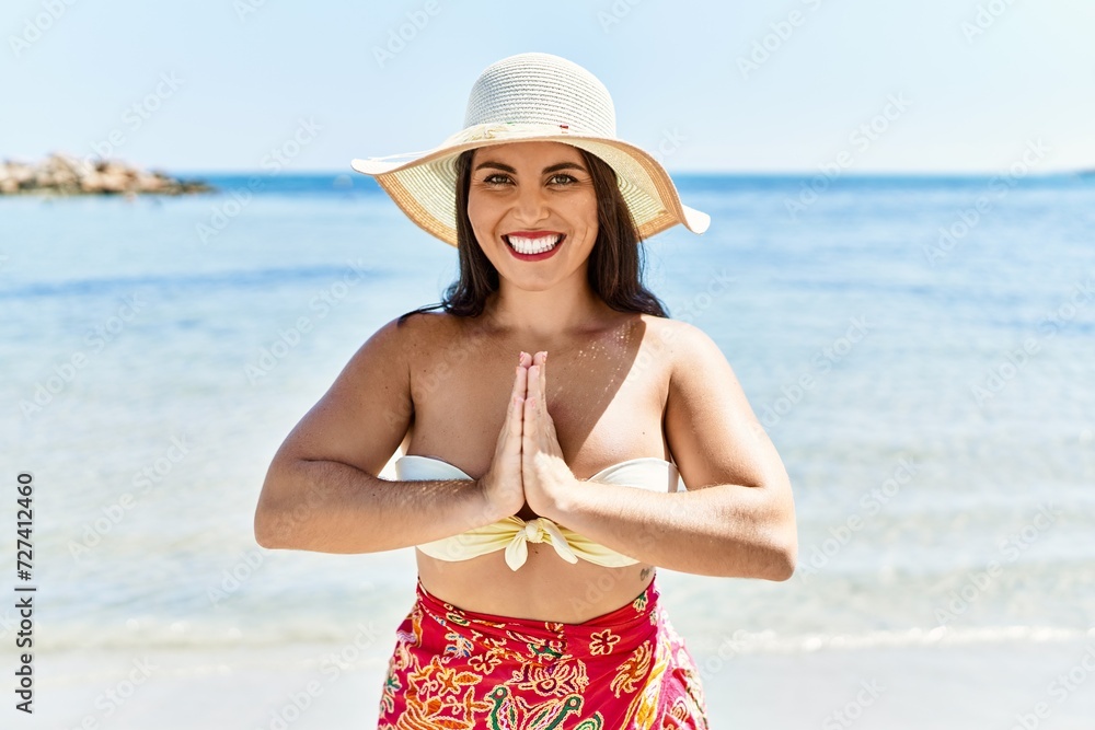 Young beautiful hispanic woman tourist wearing bikini and summer hat doing yoga exercise at seaside
