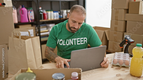 Принти на полотні Handsome young man, a dutiful volunteer, engrossed in altruistic service, working on a laptop at a community charity center, sitting amidst donation boxes