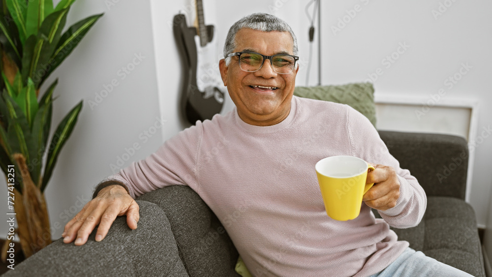 A handsome, middle-aged man with grey hair relaxes in a modern living room, smiling and holding a yellow mug. A handsome, middle-aged man with grey hair relaxes in a modern living room, smiling and holding a yellow mug.