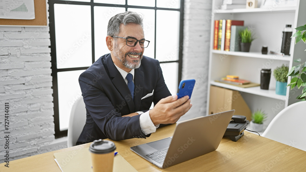 Charming young hispanic man, grey-haired employee, engages in smiling video call, effortlessly multitasking in business via laptop & smartphone in office interior Charming young hispanic man, grey-haired employee, engages in smiling video call, effortlessly multitasking in business via laptop & smartphone in office interior