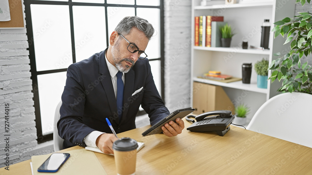 Attractive, grey-haired, young hispanic man, fully concentrated at work in his office, taking notes on touchpad, handsome professional with beard, glasses and suit sitting at table indoors Attractive, grey-haired, young hispanic man, fully concentrated at work in his office, taking notes on touchpad, handsome professional with beard, glasses and suit sitting at table indoors