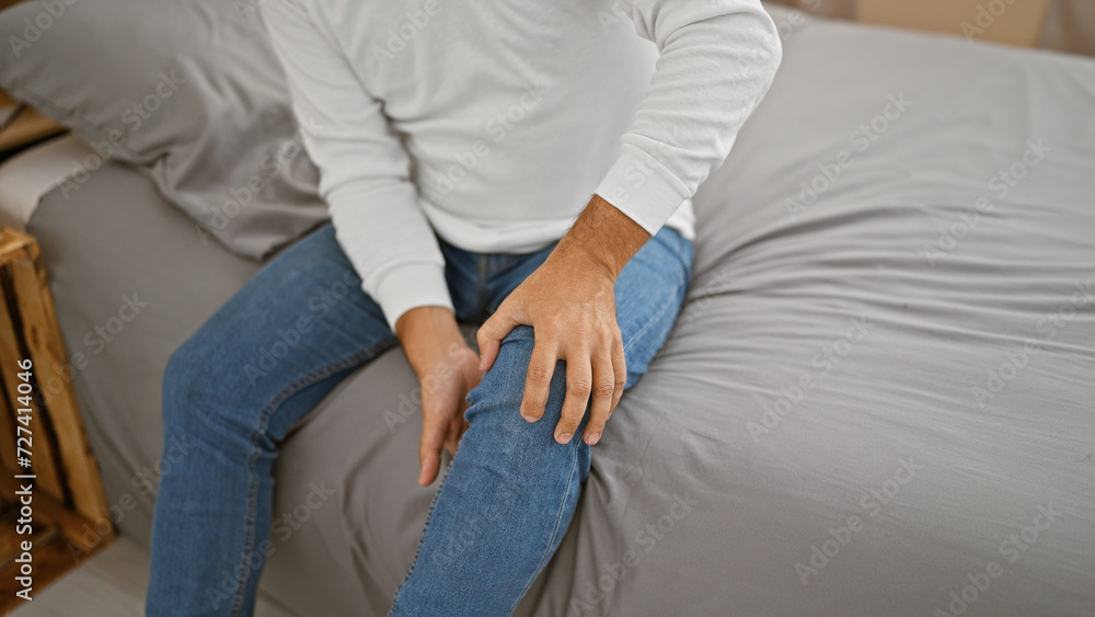 Attractive young hispanic man with grey hair, suffering from knee pain, touching his injured joint unhappily from his bed in morning. bedroom interior provides a somber indoor backdrop.
