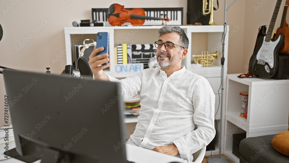 Handsome, young hispanic man, grey-haired radio news reporter making a smiling selfie with his smartphone in the lively radio studio