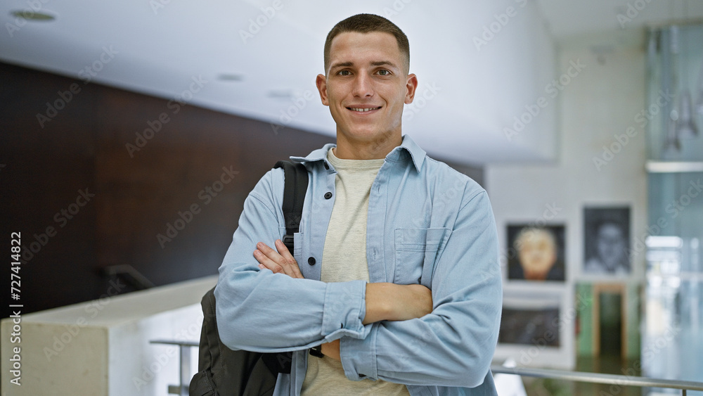 A smiling young hispanic man with crossed arms, wearing a backpack, standing in a modern university interior. A smiling young hispanic man with crossed arms, wearing a backpack, standing in a modern university interior.