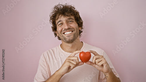 фотографія Smiling young hispanic man holding a red heart against a pink background portraying love and happiness