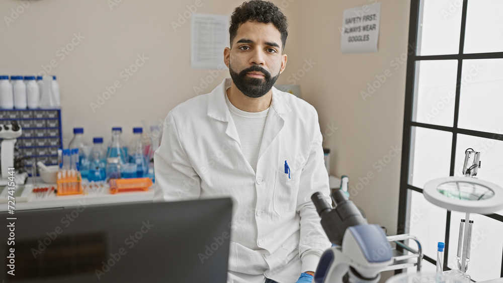 A young hispanic man with a beard, dressed in a lab coat, poses confidently in a well-equipped laboratory setting.