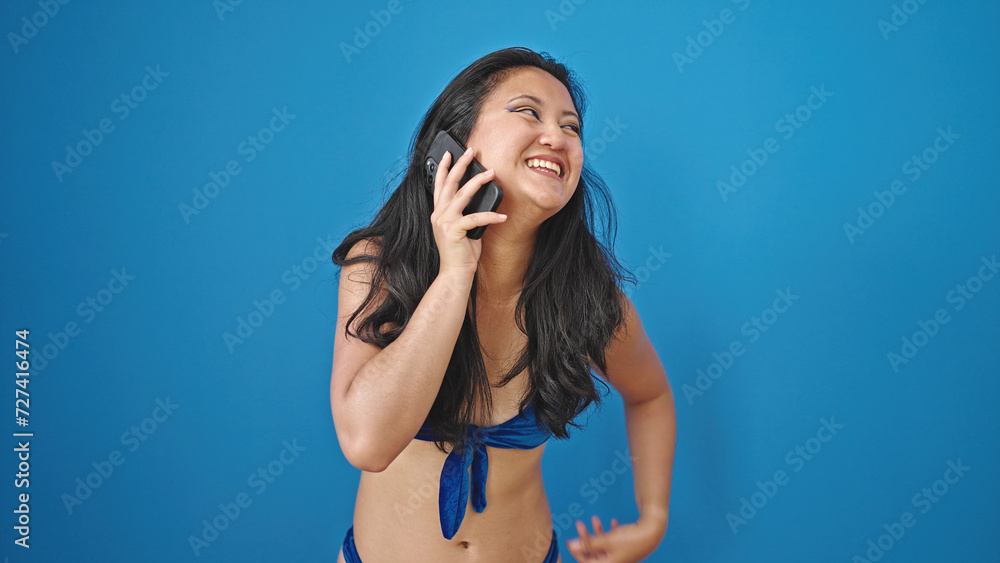 Fototapeta premium Young chinese woman tourist wearing bikini talking on smartphone over isolated blue background