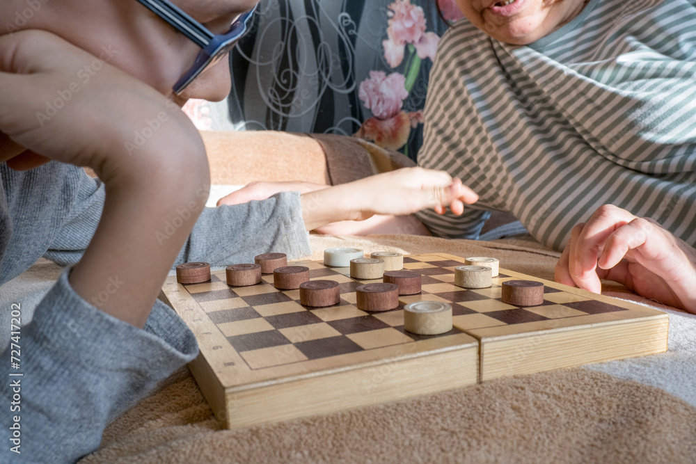 Close-up of a caring middle-aged grandmother playing checkers with a child, a boy, while lying on the bed. Happy family enjoying an interesting wooden board game at home.