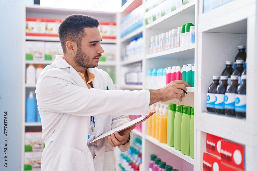 Young hispanic man pharmacist writing on document with serious expression at pharmacy