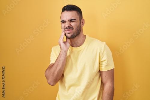 Young hispanic man standing over yellow background touching mouth with hand with painful expression because of toothache or dental illness on teeth. dentist