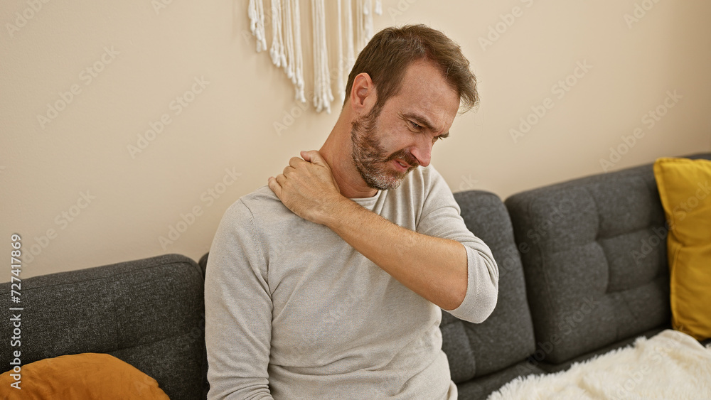 A middle-aged hispanic man with a beard grimacing in pain, touching his neck while sitting in a modern living room. A middle-aged hispanic man with a beard grimacing in pain, touching his neck while sitting in a modern living room.