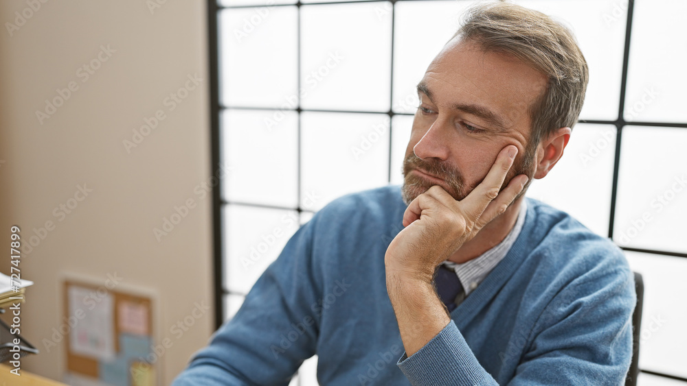 A pensive middle-aged hispanic man with a beard, wearing a sweater, rests his chin on hand in a modern office setting. A pensive middle-aged hispanic man with a beard, wearing a sweater, rests his chin on hand in a modern office setting.