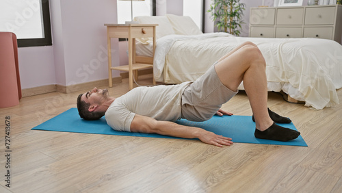 A young hispanic man exercises on a blue mat in a tidy bedroom, portraying a healthy indoor lifestyle.