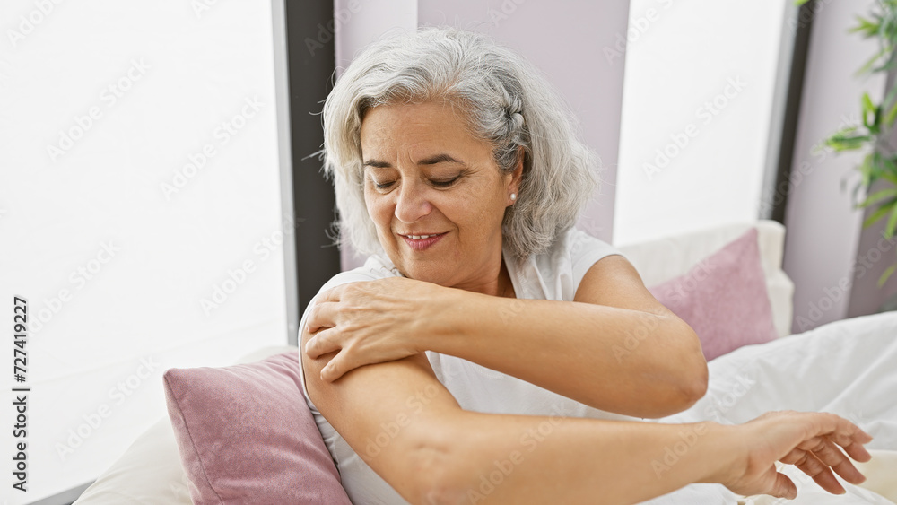 Mature woman applying lotion on her arm in a cozy bedroom setting, showcasing self-care and domestic tranquility. Mature woman applying lotion on her arm in a cozy bedroom setting, showcasing self-care and domestic tranquility.