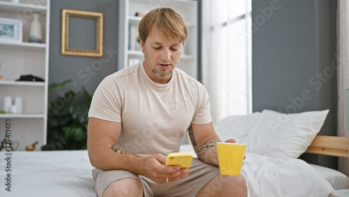 A relaxed young man with a beard checking his smartphone in a modern bedroom  holding a yellow cup  creating a casual and homely atmosphere.