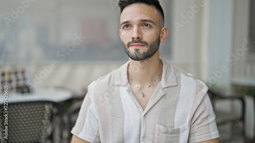 Young hispanic man sitting on bench with serious expression at coffee shop terrace