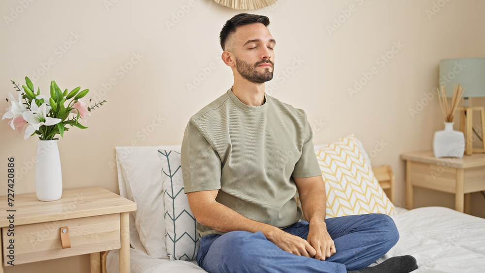 Young hispanic man sitting on bed meditating at bedroom