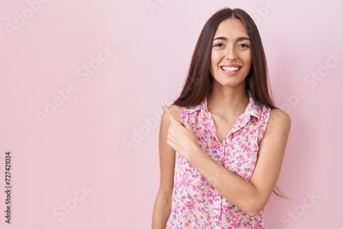 Young hispanic woman with long hair standing over pink background cheerful with a smile of face pointing with hand and finger up to the side with happy and natural expression on face