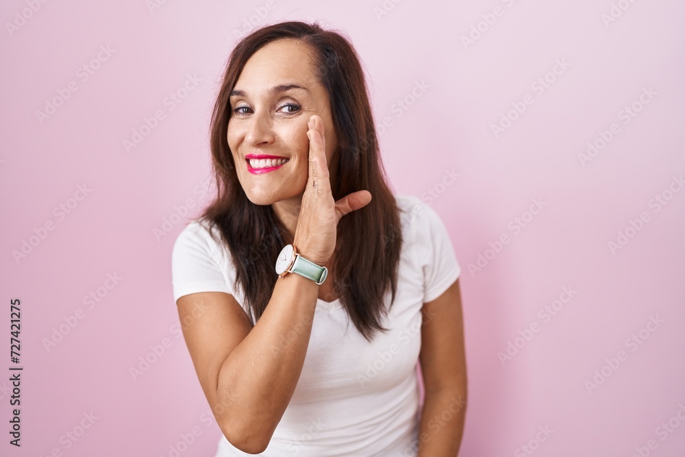 Middle age brunette woman standing over pink background hand on mouth telling secret rumor, whispering malicious talk conversation Middle age brunette woman standing over pink background hand on mouth telling secret rumor, whispering malicious talk conversation