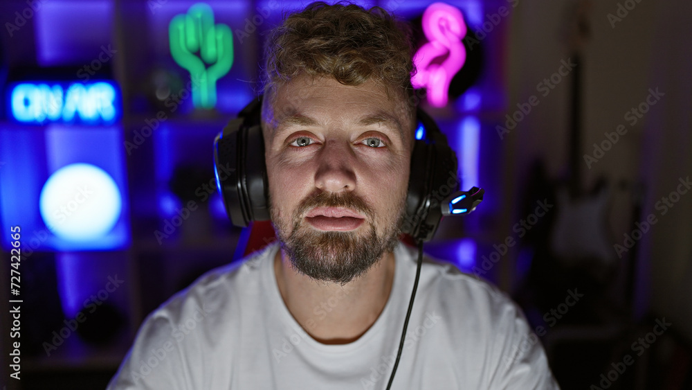 Caucasian man with blue eyes and beard wearing headphones in a dark gaming room at night Caucasian man with blue eyes and beard wearing headphones in a dark gaming room at night