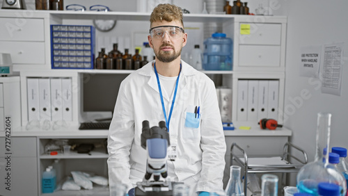 Caucasian scientist man with beard and glasses stands in laboratory surrounded by equipment and chemicals.