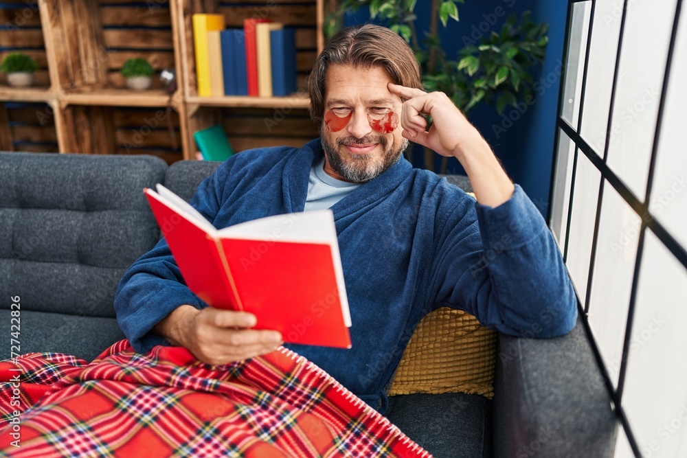 Middle age man wearing under eye patches reading book on sofa at home