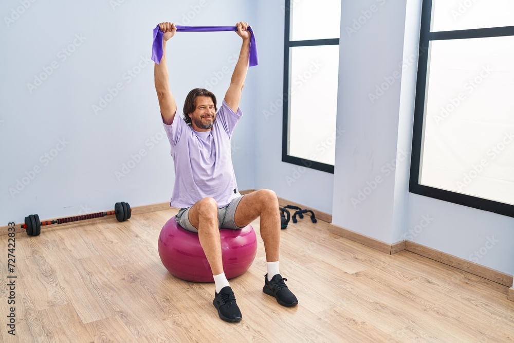 Middle age man training with elastic band sitting on fit ball at sport center Middle age man training with elastic band sitting on fit ball at sport center