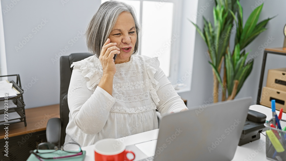 Senior woman conversing on phone with laptop in modern office