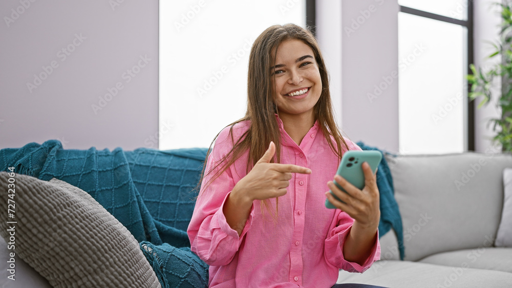 Portrait of a cheerful young hispanic woman, pointing at her smartphone while joyfully smiling and sitting on the sofa at home Portrait of a cheerful young hispanic woman, pointing at her smartphone while joyfully smiling and sitting on the sofa at home