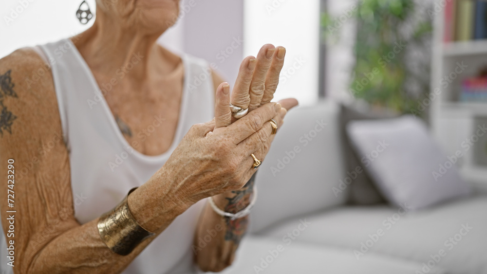 Obraz premium Senior grey-haired woman suffering intense hand pain, sitting in living room sofa at home, elderly female with short hair looking hurt, osteoarthritis problem indoors.