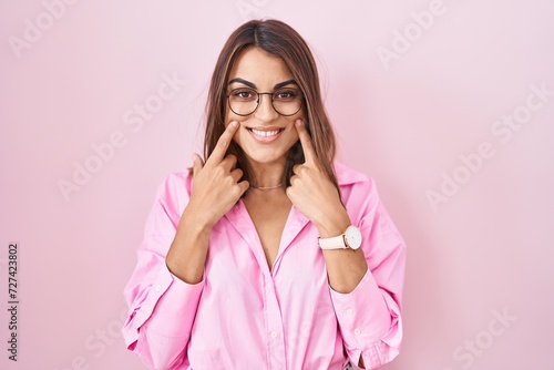 Young hispanic woman wearing glasses standing over pink background smiling with open mouth, fingers pointing and forcing cheerful smile © Krakenimages.com