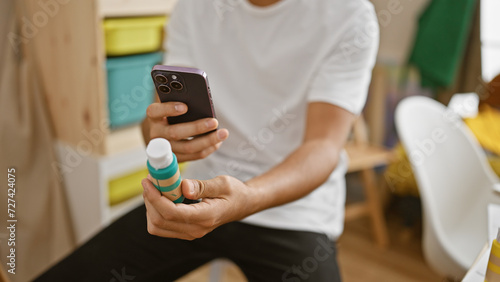 A young adult man using smartphone while holding paint bottle indoors