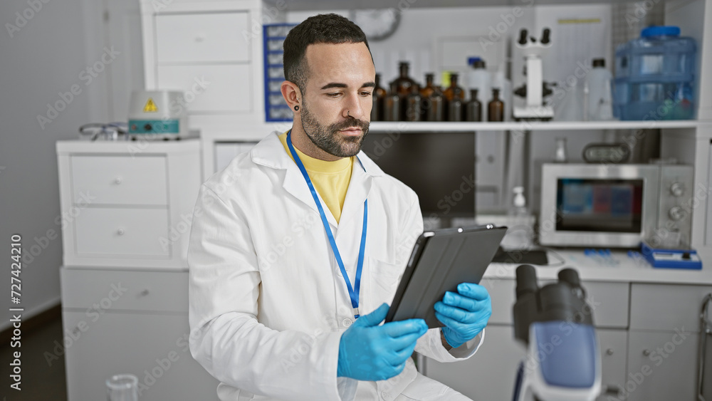 A young hispanic man with a beard wearing a lab coat and gloves holds a tablet in a medical laboratory setting. A young hispanic man with a beard wearing a lab coat and gloves holds a tablet in a medical laboratory setting.
