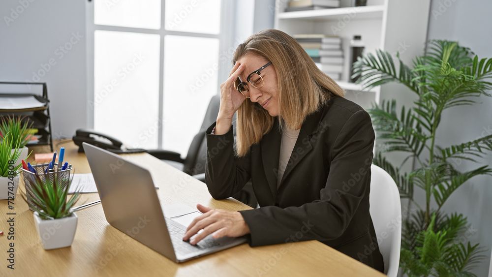 Exhausted young businesswoman with blonde hair suffering from a severe migraine at work, struggling to focus on her laptop, in the office atmosphere.