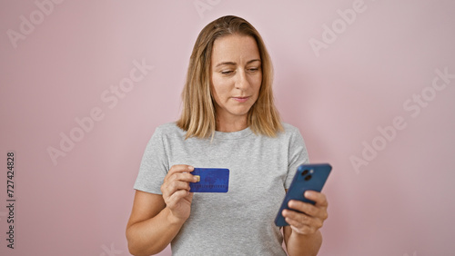 Blonde adult texting a serious payment message over pink isolated background. young attractive female strains with credit card, cellphone issues and unwelcome financial investment problem. photo