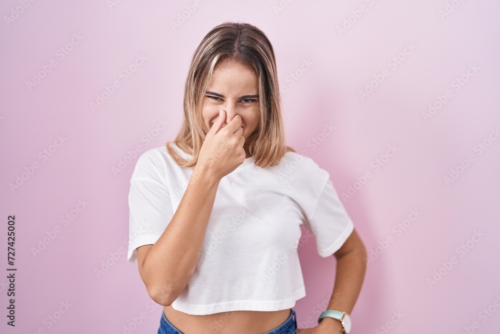 Young blonde woman standing over pink background smelling something stinky and disgusting, intolerable smell, holding breath with fingers on nose. bad smell Young blonde woman standing over pink background smelling something stinky and disgusting, intolerable smell, holding breath with fingers on nose. bad smell