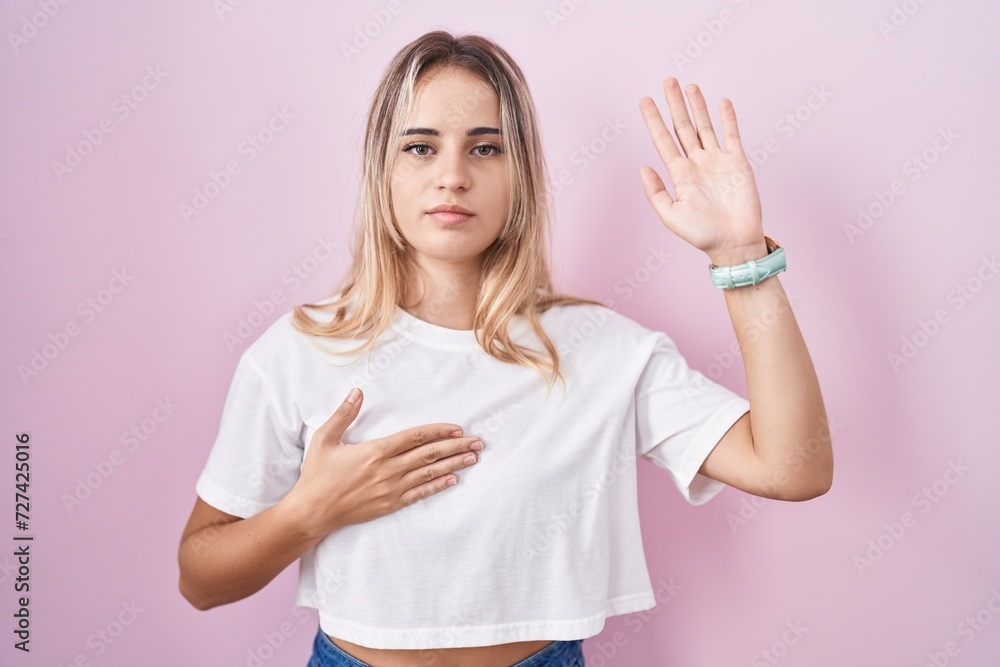 Young blonde woman standing over pink background swearing with hand on chest and open palm, making a loyalty promise oath Young blonde woman standing over pink background swearing with hand on chest and open palm, making a loyalty promise oath