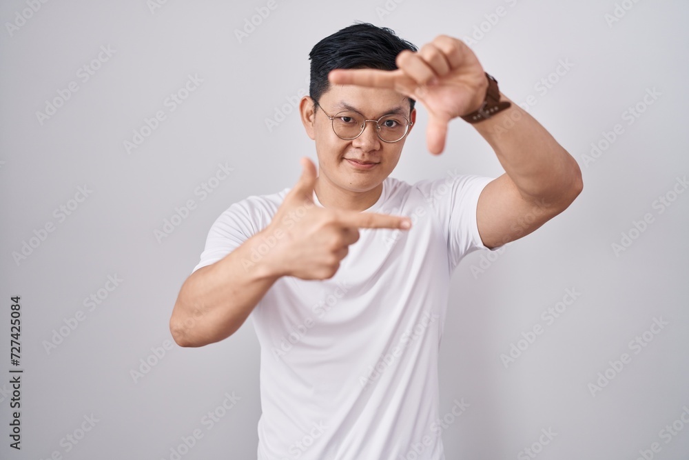 Naklejka premium Young asian man standing over white background smiling making frame with hands and fingers with happy face. creativity and photography concept.
