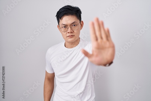 Young asian man standing over white background doing stop sing with palm of the hand. warning expression with negative and serious gesture on the face.