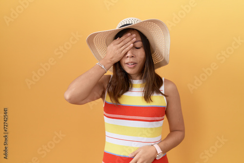 Middle age chinese woman wearing summer hat over yellow background yawning tired covering half face, eye and mouth with hand. face hurts in pain.