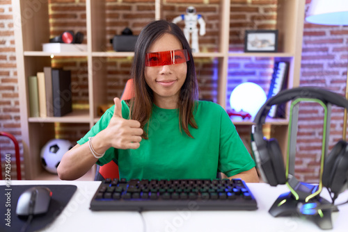 Middle age chinese woman wearing virtual reality glasses doing happy thumbs up gesture with hand. approving expression looking at the camera showing success.