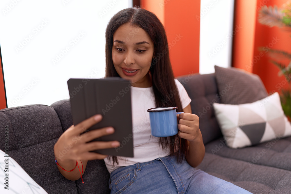 Young arab woman using touchpad drinking coffee sitting on sofa at home Young arab woman using touchpad drinking coffee sitting on sofa at home