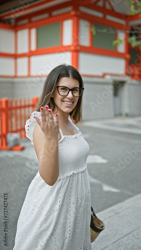 Cheerful and beautiful hispanic woman in glasses invites with friendly gesture to a japanese temple in tokyo, japan