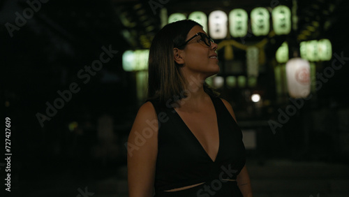 Cheerful beautiful hispanic woman in glasses enjoying kyoto's nightlife, looking around and smiling confidently on a lighted japanese street