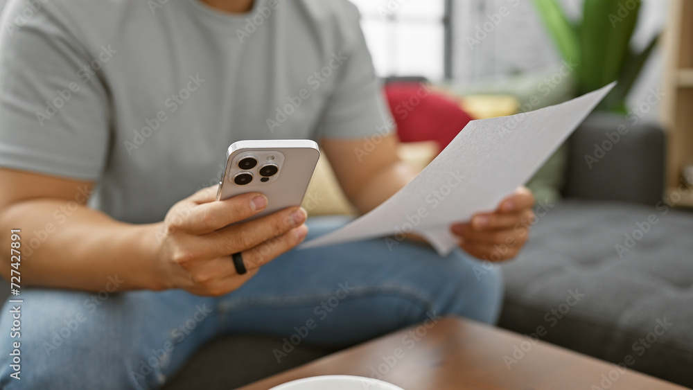 A young asian man checks a document while holding a smartphone in a modern living room setting. A young asian man checks a document while holding a smartphone in a modern living room setting.