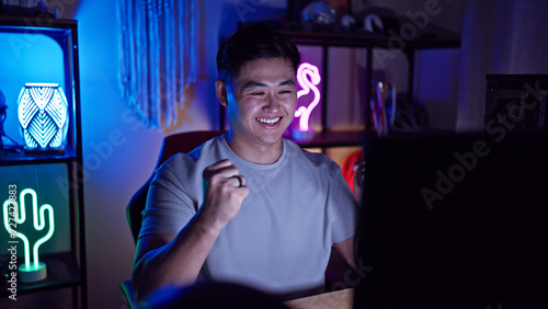 A cheerful asian man enjoys gaming in a dark room illuminated by neon lights, exuding a cozy, tech-savvy atmosphere.