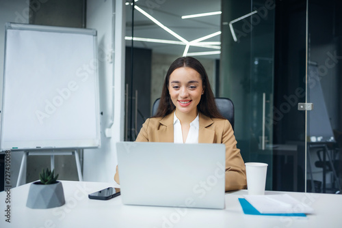 Portrait of a cheerful businesswoman sitting at the table in office look at laptop work on project
