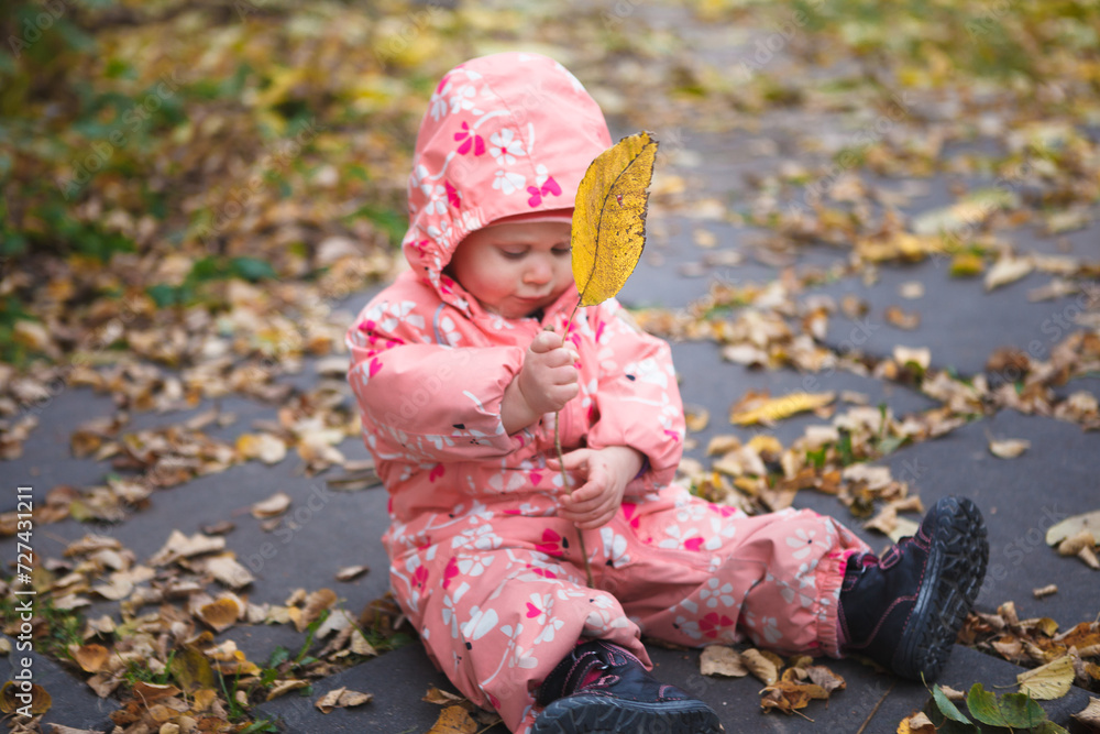A little girl in a peach jumpsuit is sitting on the ground and playing with yellow autumnal leaf. 