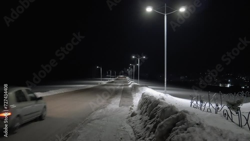 A small car passing on a snow-covered road during a winter night. Street poles light the way. City lights visible in the distance. Handheld shot photo