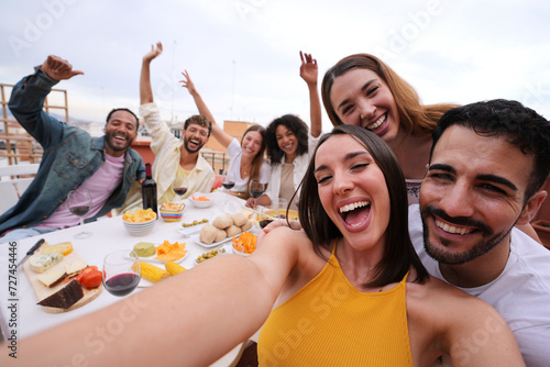 Excited young pretty woman taking group selfie with joyful friends on rooftop. Cheerful people poses making smiling faces to camera outdoor gathered sitting table at lunch raising happy arms for photo photo